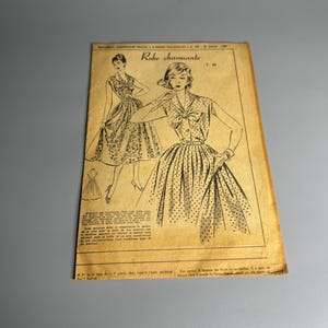 May include: A black and white illustration of a vintage dress pattern from a French magazine. The pattern is called "Robe charmante" and features a dress with a bow at the neck and a skirt with a polka dot pattern.