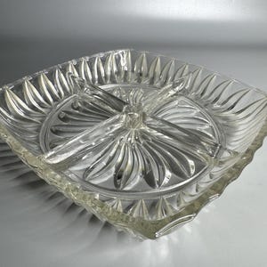 May include: Clear glass divided serving dish with a square shape and a central circular section. The dish has four compartments and a decorative, ridged design radiating from the center. The edges are scalloped.