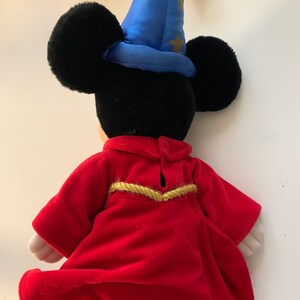 Może przedstawiać: Pluszowa zabawka Mickey Mouse w czerwonej szacie z złotymi ozdobami i niebieskim kapeluszu czarodzieja z złotymi gwiazdami.