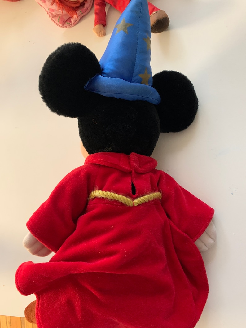 Może przedstawiać: Pluszowa zabawka Mickey Mouse w czerwonej szacie z złotymi ozdobami i niebieskim kapeluszu czarodzieja z złotymi gwiazdami.