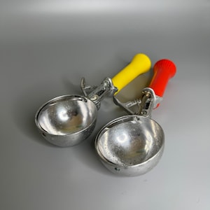May include: Two ice cream scoops with yellow and red handles. The scoops are made of metal and have a shiny finish.