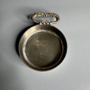 May include: A round silver-toned metal dish featuring a decorative handle shaped like two opposing dragon heads. The interior base is engraved with the text CONSERVES BEG-AVEL LOUIS MARC BREST. The metal surface shows a weathered, aged patina.