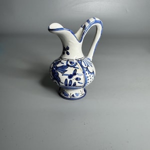 May include: A small, white ceramic pitcher with a blue floral design. The pitcher has a curved handle and a spout. The base and handle are decorated with blue stripes. The pitcher is approximately 6 inches tall.