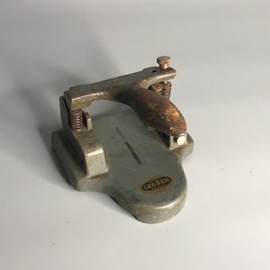 Peut inclure: Un perforateur de papier vintage en métal gris avec une poignée en bois. Le perforateur porte une étiquette indiquant "Machine Leober Depuis 1901".