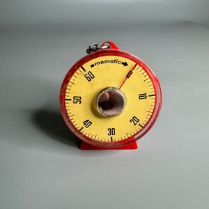 May include: A round red Mematic kitchen timer with a pale yellow face featuring black numerals from 10 to 60. The device has a clear plastic cover, a central metallic knob, and a small metal loop at the top for hanging or carrying.