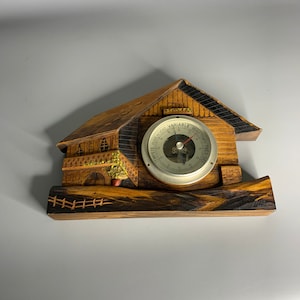 May include: A wooden barometer shaped like a house with a carved wooden fence and a small tree. The barometer face is round and silver with black markings and a red needle. The face reads "VARIABLE REALTIME TEMP."