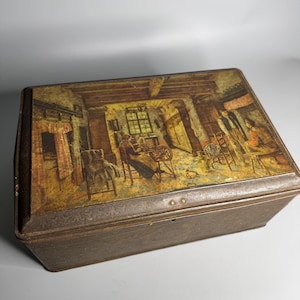 May include: A vintage tin box with a painted scene of a room with people inside. The box is brown with gold accents and has a hinged lid.