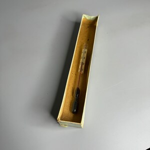 May include: A vintage glass thermometer with a black bulb, resting inside a worn, rectangular cardboard box. The thermometer has a red liquid column and temperature markings. The box is beige with a slightly distressed appearance.