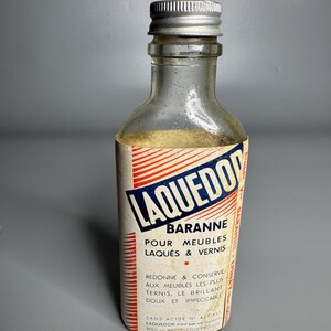 May include: A small, clear glass bottle with a silver cap. The bottle has a label with red, white, and blue text that reads "LAQUEDOR BARANNE POUR MEUBLES LAQUÉS & VERNIS". The bottle appears to be vintage.