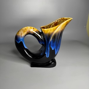 Puede incluir: Una jarra de cerámica negra con un esmalte azul y marrón. La jarra tiene un diseño único y curvo y una abertura ancha.