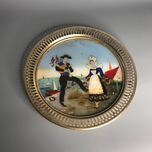 Może przedstawiać: Złota metalowa taca z ozdobną, ażurową krawędzią. Taca przedstawia namalowaną scenę przedstawiającą mężczyznę i kobietę w tradycyjnych strojach stojących na molo z żaglówkami w tle.