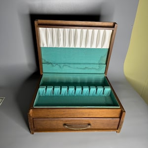 May include: Open wooden silverware chest with a drawer. The interior is lined with teal fabric and has slots for utensils. The top section is lined with white pleated fabric. The chest has a brass-colored handle on the drawer.