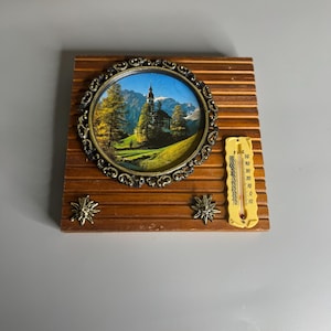 May include: A vintage wooden wall decor piece featuring a round framed image of a church in a mountain landscape. The frame is ornate, and the piece includes a thermometer and two decorative star-shaped accents.