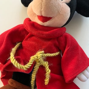 Może przedstawiać: Pluszowa zabawka Mickey Mouse ubrana w czerwoną szatę z pasem ze złotej liny i niebieskim kapeluszem.