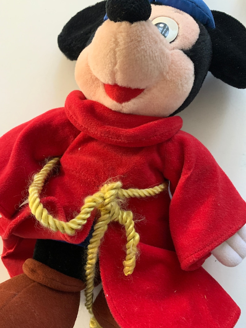 Może przedstawiać: Pluszowa zabawka Mickey Mouse ubrana w czerwoną szatę z pasem ze złotej liny i niebieskim kapeluszem.