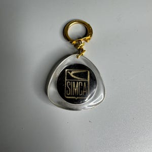 May include: A vintage Simca keychain with a gold-colored ring and a clear, triangular pendant. The pendant features a black and gold Simca logo. The keychain is likely from the automotive industry.