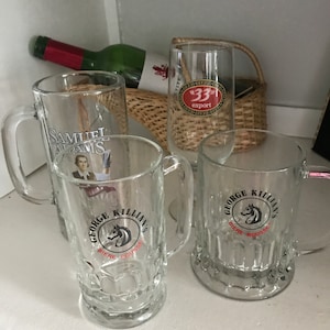 May include: Clear glass beer mugs with handles, featuring the "George Killian's Biere Rousse" logo and a Samuel Adams mug. A green wine bottle and a tall glass with a "33 Export" label are also visible.