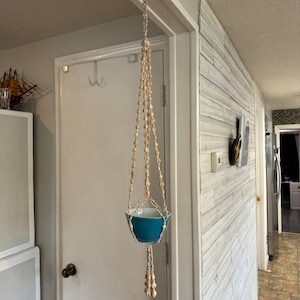 Może przedstawiać: Niebieska ceramiczna doniczka wisząca na makramowym hamakach dla roślin wykonanym z naturalnych beżowych koralików. Hamak jest zawieszony na haczyku na białym suficie.
