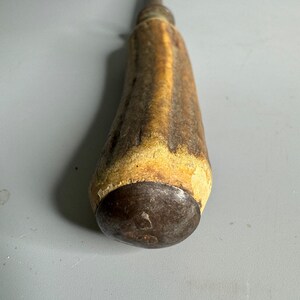 May include: A vintage, metal knife sharpener with a brown antler handle. The handle has a worn, rustic look. The metal tip is rounded and has a small, circular mark.