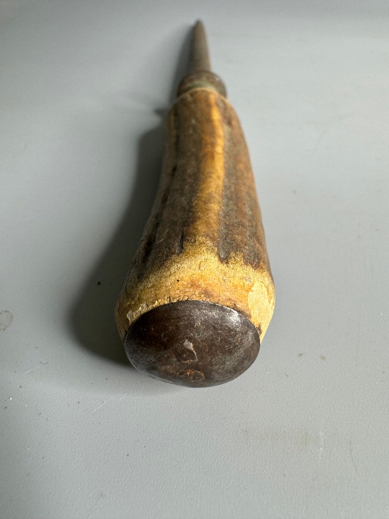 May include: A vintage, metal knife sharpener with a brown antler handle. The handle has a worn, rustic look. The metal tip is rounded and has a small, circular mark.