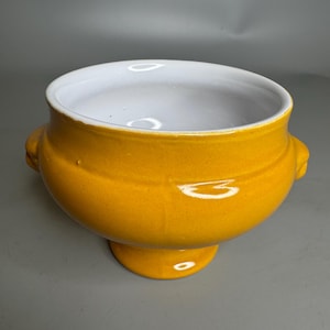 Puede incluir: Una sopera de cerámica amarilla con interior blanco y dos asas. La sopera tiene un cuerpo redondo y una base de pedestal.