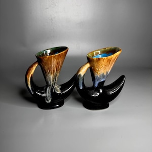 Puede incluir: Dos tazas de cerámica con un diseño único. Las tazas tienen una base y un asa negras, con una sección superior marrón y beige. El interior de las tazas es azul. Las tazas son probablemente de estilo vintage o retro.