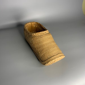 Puede incluir: Una cesta tejida con forma de zapato, hecha de paja natural de color marrón claro.