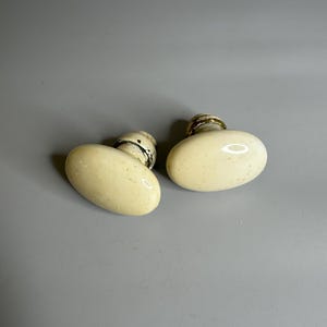 Puede incluir: Dos pomos de puerta ovalados de color marfil antiguo con bases de metal envejecido. Los pomos tienen una superficie lisa, ligeramente moteada y una estética clásica y vintage. Se encuentran sobre un fondo gris liso.