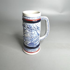 May include: Tall ceramic beer stein with a white rim and handle, featuring blue and white relief artwork of airplanes and clouds. The stein has a cylindrical shape with a red and blue band near the top and bottom. The handle is decorated with a blue and white pattern.