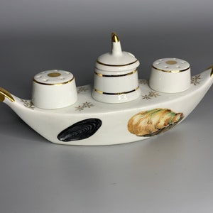 May include: White ceramic salt and pepper shaker set shaped like a boat with gold accents. The boat has a small, round, white ceramic container in the center with a lid. The boat is decorated with black and brown seashells.