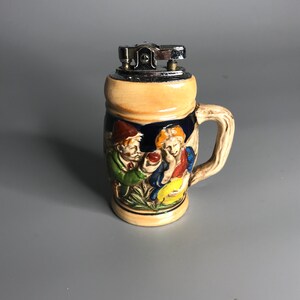 Könnte beinhalten: Ein Keramik-Bierkrug in Form eines Feuerzeugs mit einem blauen und weißen Design, das drei Personen in einer Szene zeigt. Das Feuerzeug hat ein silbernes Oberteil.