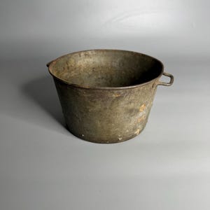 May include: A weathered, antique metal bucket with a slightly tapered shape. The bucket has a dark, aged appearance with visible rust and wear, and a small handle on one side. The overall color is a muted, greenish-brown.