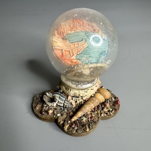 May include: A glass snow globe with a seashell scene inside. The globe is on a base made of seashells and sand.