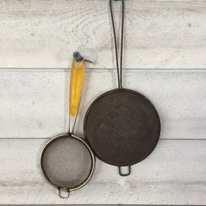 May include: Two metal strainers hanging on a white wooden wall. The larger strainer has a dark metal finish and a wire handle. The smaller strainer has a yellow plastic handle and a silver finish.