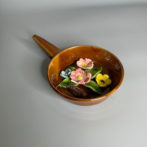 May include: A brown ceramic dish with a handle, featuring a floral design with pink, yellow, blue, and green flowers.