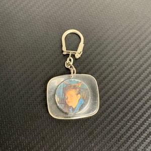 May include: A clear, square-shaped plastic keychain featuring a circular insert with a printed portrait of a person with dark hair wearing a blue jacket. The keychain includes a silver-toned metal split ring attached by a small chain link.