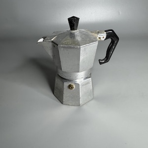 May include: A silver aluminium stovetop espresso maker with a black handle and a black top. The coffee pot has a hexagonal base and a triangular spout. The handle is curved and attached to the side of the pot.