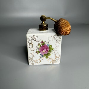 May include: A white porcelain perfume bottle with a gold floral design and a brown wicker atomizer. The bottle has a square shape and a floral design with a pink rose.