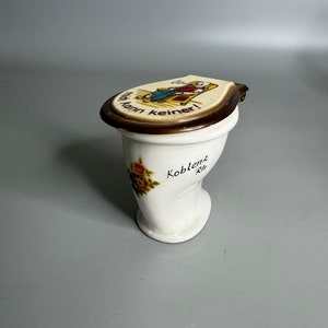 Op de afbeelding: Witte keramische spaarpot in de vorm van een toilet met een bruin deksel. Het deksel heeft een cartoonafbeelding van een persoon die op een toilet zit met de tekst "Hier kann keiner!" De basis van de spaarpot heeft een bloemenmotief en de tekst "Koblenz Rh."
