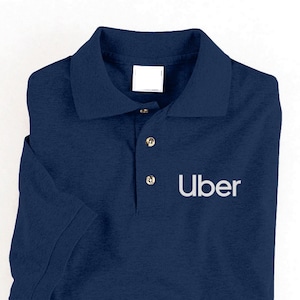 May include: Navy blue polo shirt with short sleeves and a collared neckline. The shirt features the word "Uber" in white block letters on the front. The shirt is folded neatly and has three gold-colored buttons.