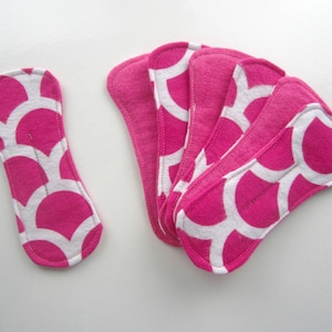 Puede incluir: Compilación de compresas menstruales reutilizables en rosa y blanco con un diseño de vieira. Las compresas están hechas de tela suave y están diseñadas para la comodidad y la absorción. La imagen muestra una compresa individual y un grupo de cuatro.