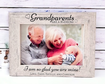 May include: A white wooden picture frame with a photo of grandparents holding a baby. The frame has the text "Grandparents are a blessing" and "I am so glad you are mine! Love, Theo, Estelle, and Cameron."