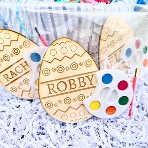 May include: Two wooden Easter eggs with the names "Robby" and "Rach" painted on them. The eggs are surrounded by white shredded paper and a small white paint palette with six paint wells.