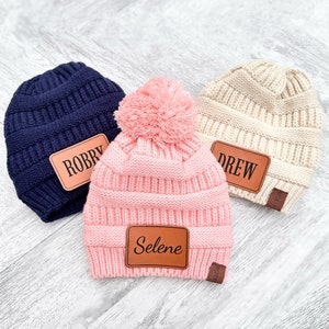 May include: Three knitted beanies with pom poms and personalized leather patches. The beanies are navy blue, pink, and beige. The patches read "Robby", "Selene", and "Drew".