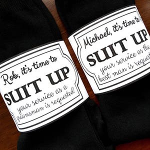 K&ouml;nnte beinhalten: Zwei schwarze Socken mit wei&szlig;en Etiketten, die daran befestigt sind. Die Etiketten haben schwarzen Text, der lautet: "Rob, es ist Zeit, sich anzuziehen, dein Dienst als Trauzeuge ist erforderlich!" und "Michael, es ist Zeit, sich anzuziehen, dein Dienst als Trauzeuge ist erforderlich!"