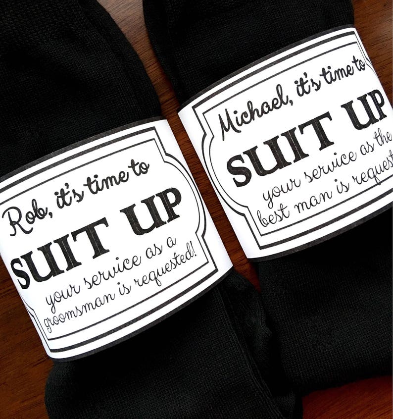 K&ouml;nnte beinhalten: Zwei schwarze Socken mit wei&szlig;en Etiketten, die daran befestigt sind. Die Etiketten haben schwarzen Text, der lautet: "Rob, es ist Zeit, sich anzuziehen, dein Dienst als Trauzeuge ist erforderlich!" und "Michael, es ist Zeit, sich anzuziehen, dein Dienst als Trauzeuge ist erforderlich!"