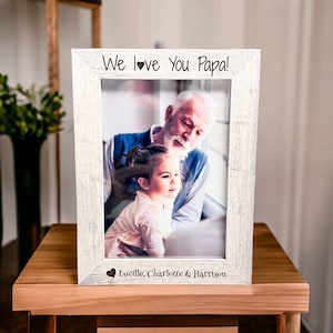 May include: A white wooden picture frame with a photo of an older man and a young girl. The frame has the text "We love you Papa!" and "Lucille, Charlotte & Harrison" engraved on it.