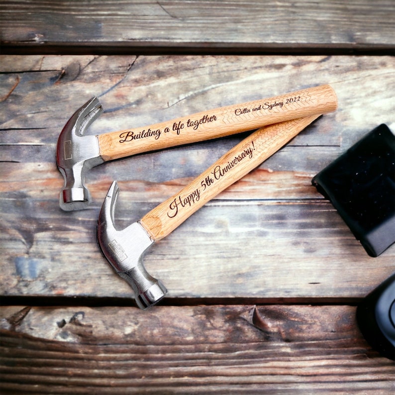 Op de afbeelding: Twee houten hamers met zilveren koppen, gegraveerd met de tekst "Building a life together Ollie and Sydney 2022" en "Happy 5th Anniversary!" De hamers liggen op een houten oppervlak.