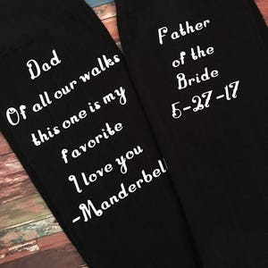 May include: A pair of black socks with white text. One sock reads "Dad Of all our walks this one is my favorite I love you -Manderbel". The other sock reads "Father of the Bride 5-27-17".
