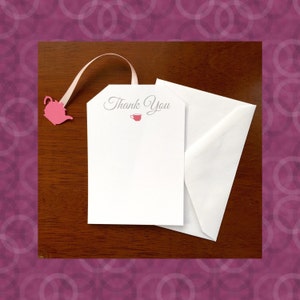 May include: A white card with the words "Thank You" in gray script, a small red teacup icon, and a pink teapot-shaped tag attached with a ribbon. The card is next to a white envelope on a dark wood surface.